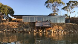 Een modern huis met een glazen gevel, een terras en uitzicht op een wateroppervlak.
