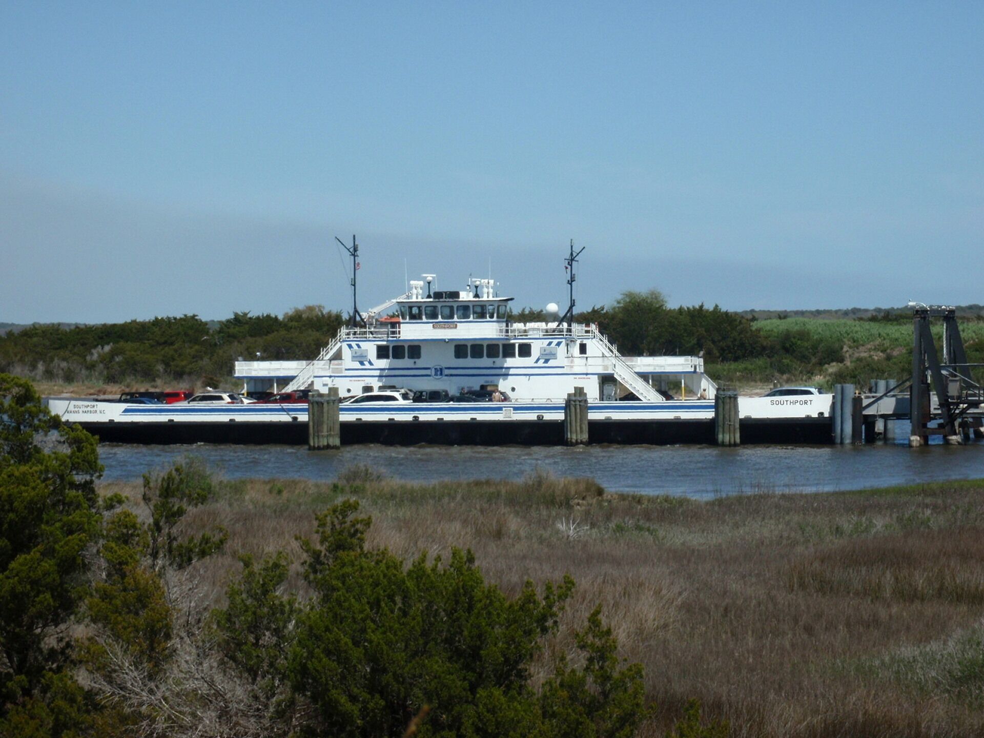A large white ferry with blue stripes docked at a pier, surrounded by grassy areas and trees.