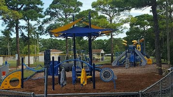 Explore a playground with a climbing frame, slide, and swing set, surrounded by a fence and shaded by trees.