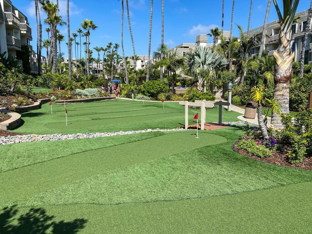 Een golfbaan met een putting green, omringd door palmbomen en een gebouw met balkons.