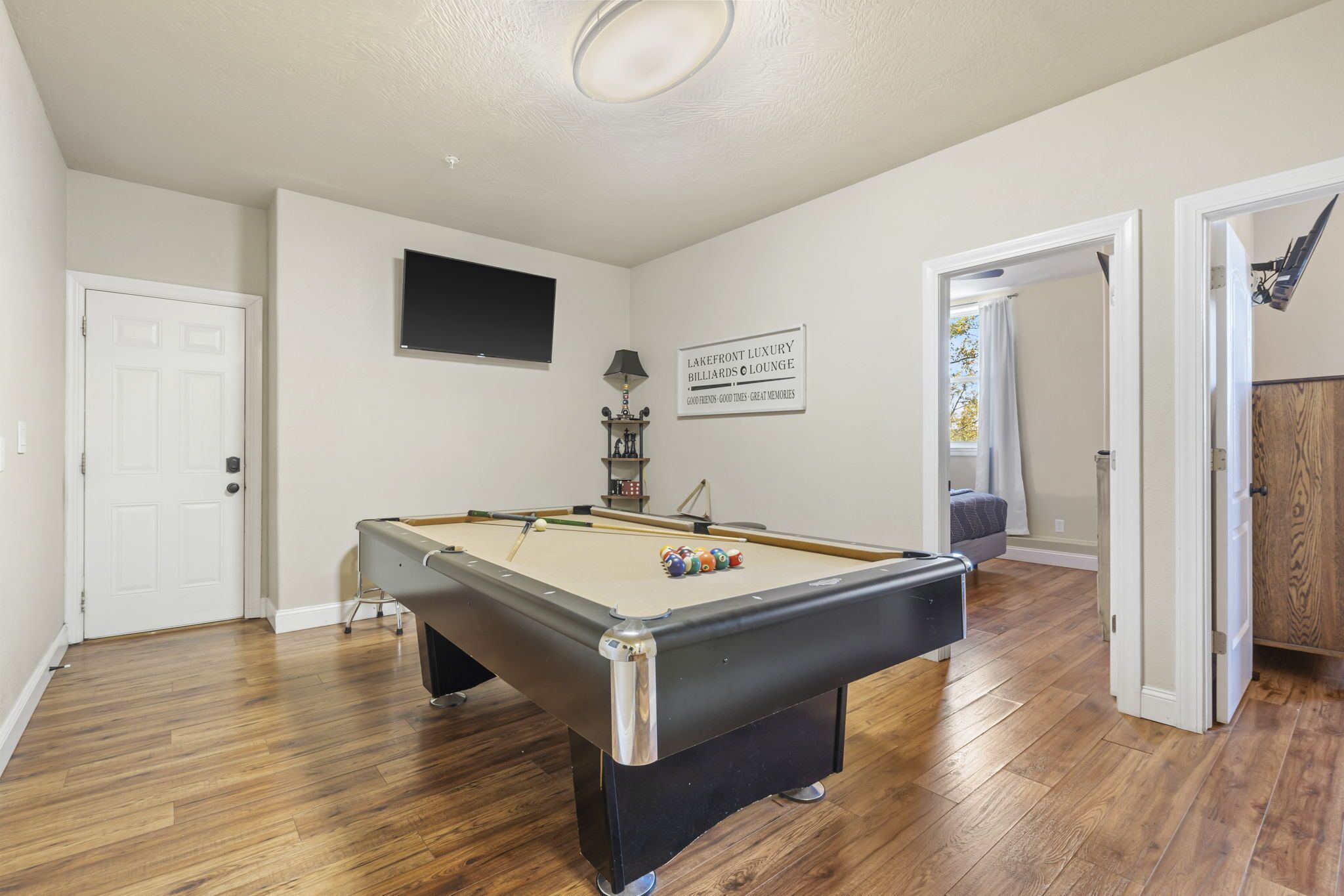 A pool table in a room with wooden flooring, a door, a television mounted on the wall, and a sign above the table.