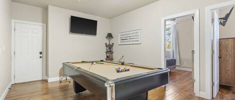 A pool table in a room with wooden flooring, a door, a television mounted on the wall, and a sign above the table.
