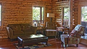 L’intérieur d’une cabane en rondins, avec un canapé, un fauteuil et un pouf, un plancher en bois et de grandes fenêtres.