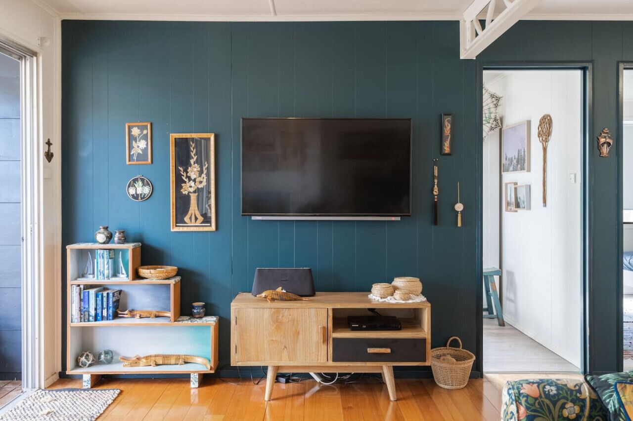 A living room with a large flat-screen TV mounted on the wall, wooden shelves with various decorative items, a wooden cabinet with a laptop, and a wicker basket.