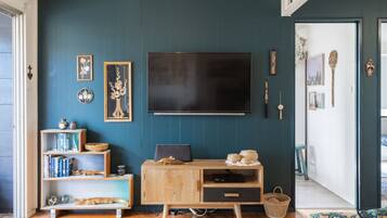 A living room with a large flat-screen TV mounted on the wall, wooden shelves with various decorative items, a wooden cabinet with a laptop, and a wicker basket.