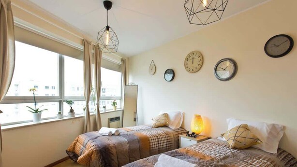 Een hotelkamer met twee bedden, een raam met gordijnen, meerdere klokken aan de muur en een lampje op het nachtkastje.