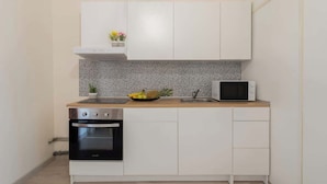 Une cuisine moderne avec des meubles blancs, un four en acier inoxydable, un micro-ondes et un petit plan de travail agrémenté d’un panier de fruits et d’une plante en pot.