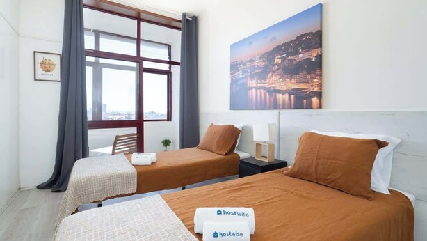 Une chambre d’hôtel avec deux lits, une grande fenêtre, un tableau représentant un paysage urbain et une pancarte sur laquelle on peut lire « hostwise ».