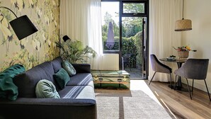 Ein modernes Wohnzimmer mit einer Couch, einem Esstisch mit Stühlen, einem großen Fenster mit Vorhängen und Blick ins Grüne.