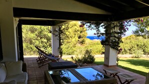 A covered outdoor seating area with wicker furniture and a glass-top table, overlooking a garden and the sea. - Big beautiful villa sleeps 10: sea view, beach 5 minutes and everything nearby (Es Mercadal)