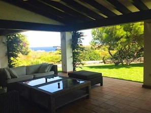 A covered patio with a sofa and a glass-top coffee table, overlooking a lush garden and a distant view of the sea.