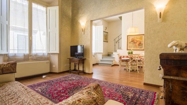 A spacious living room with a large window, a television, a patterned rug, and a staircase leading to another room. - Tornaquinci Luxury Penthouse (Firenze)