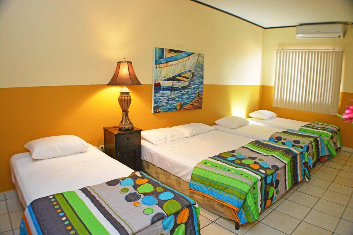 A hotel room with four single beds, a nightstand with a lamp, a painting of a boat on the wall, and a window with blinds.