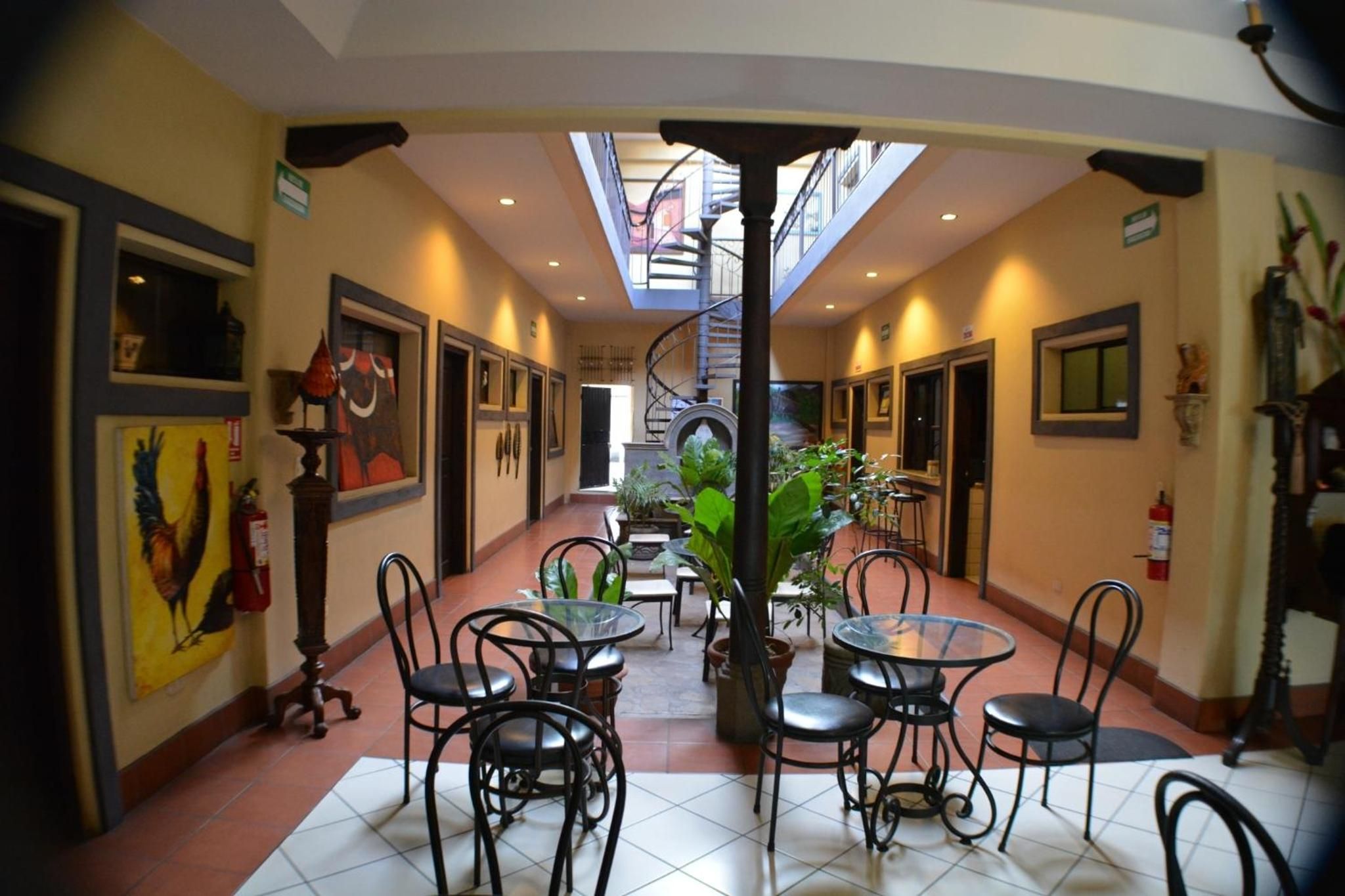 A hotel corridor with a central staircase, multiple dining tables and chairs, and potted plants.