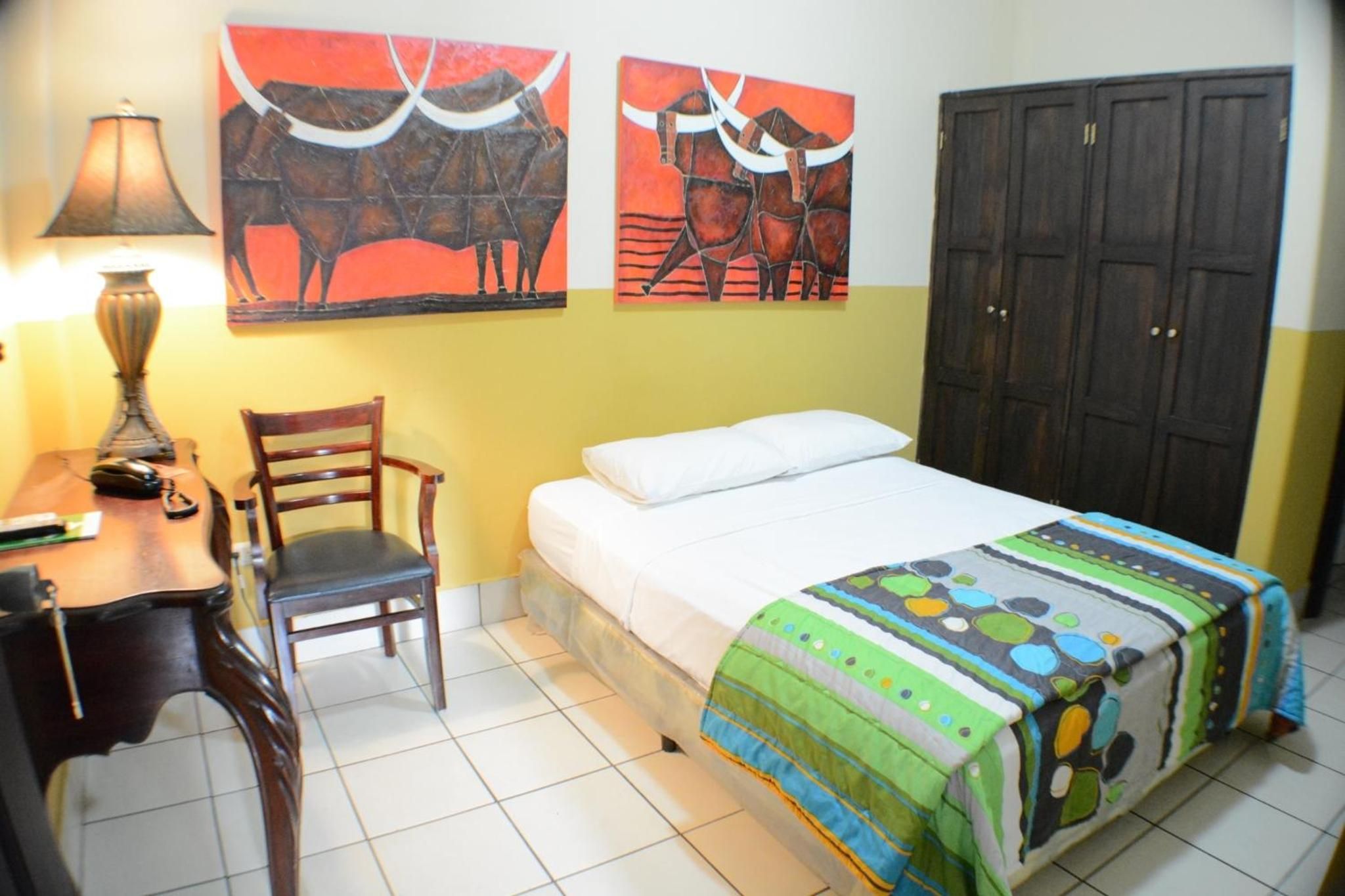 Un dormitorio con una cama, una silla, un escritorio, una lámpara y dos cuadros en la pared.