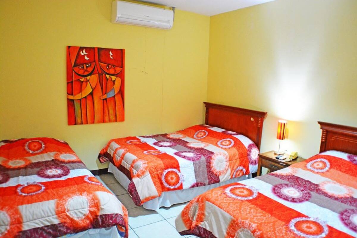 Une chambre d’hôtel avec trois lits, chacun recouvert d’une couvre-lit à motifs. Il y a une tête de lit en bois sur le côté droit, un climatiseur fixé au mur et un tableau au-dessus des lits.