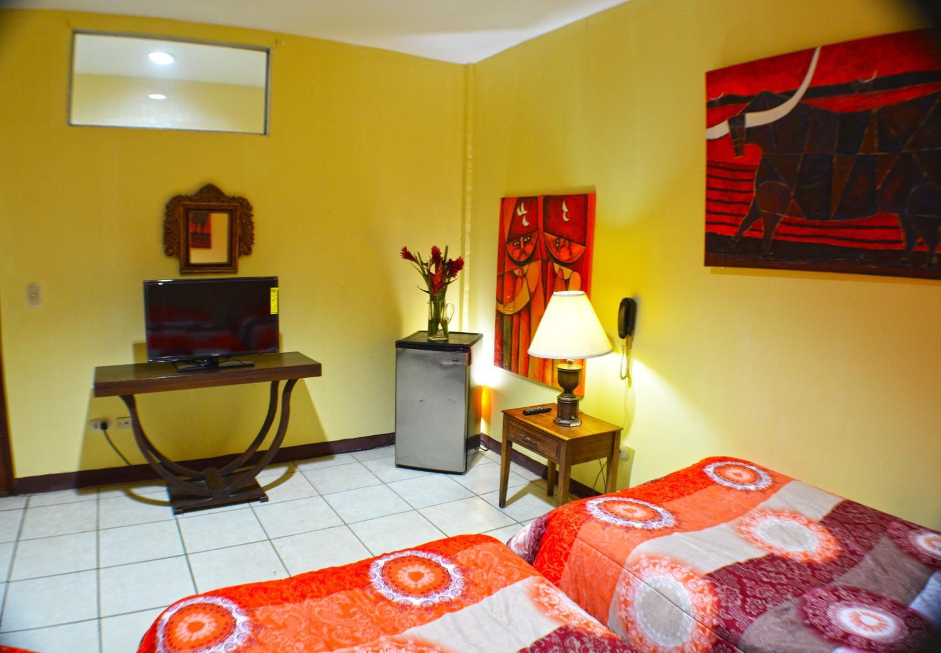Habitación de hotel con cama, escritorio, televisor, refrigerador, lámpara y un cuadro en la pared.