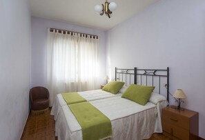 A double bed with green pillows, a bedside table with a lamp, a chair, and a window with curtains.