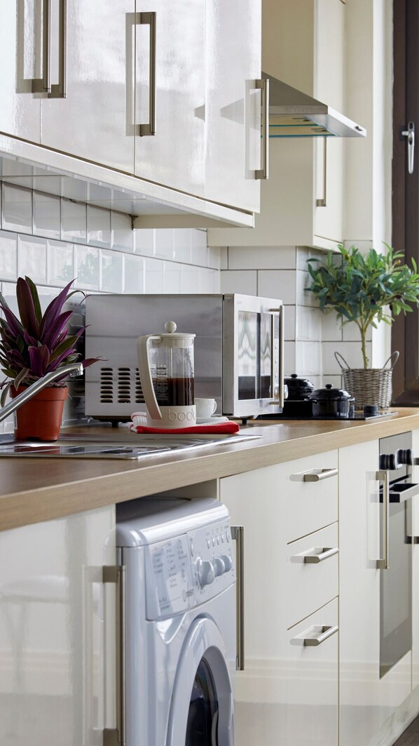 A modern kitchen with white cabinets, a stainless steel washing machine, a coffee maker, and potted plants.