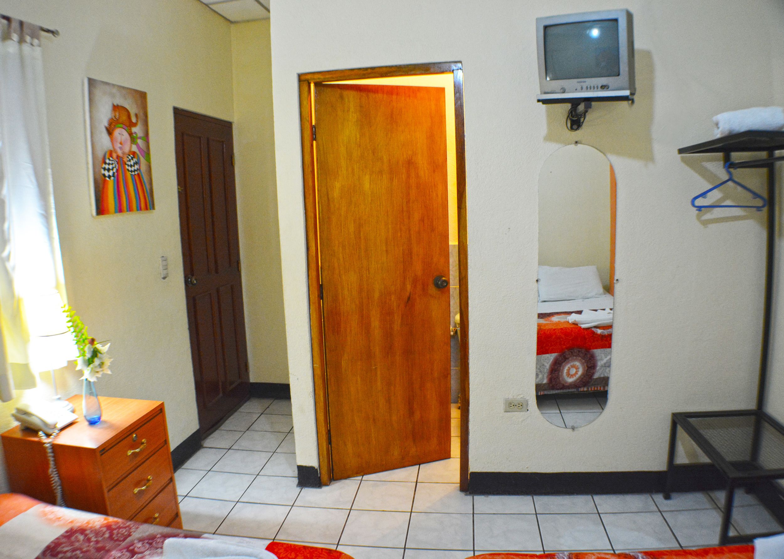 Habitación de hotel con puerta de madera, cama, una mesita, televisor montado en la pared y un cuadro en la pared.
