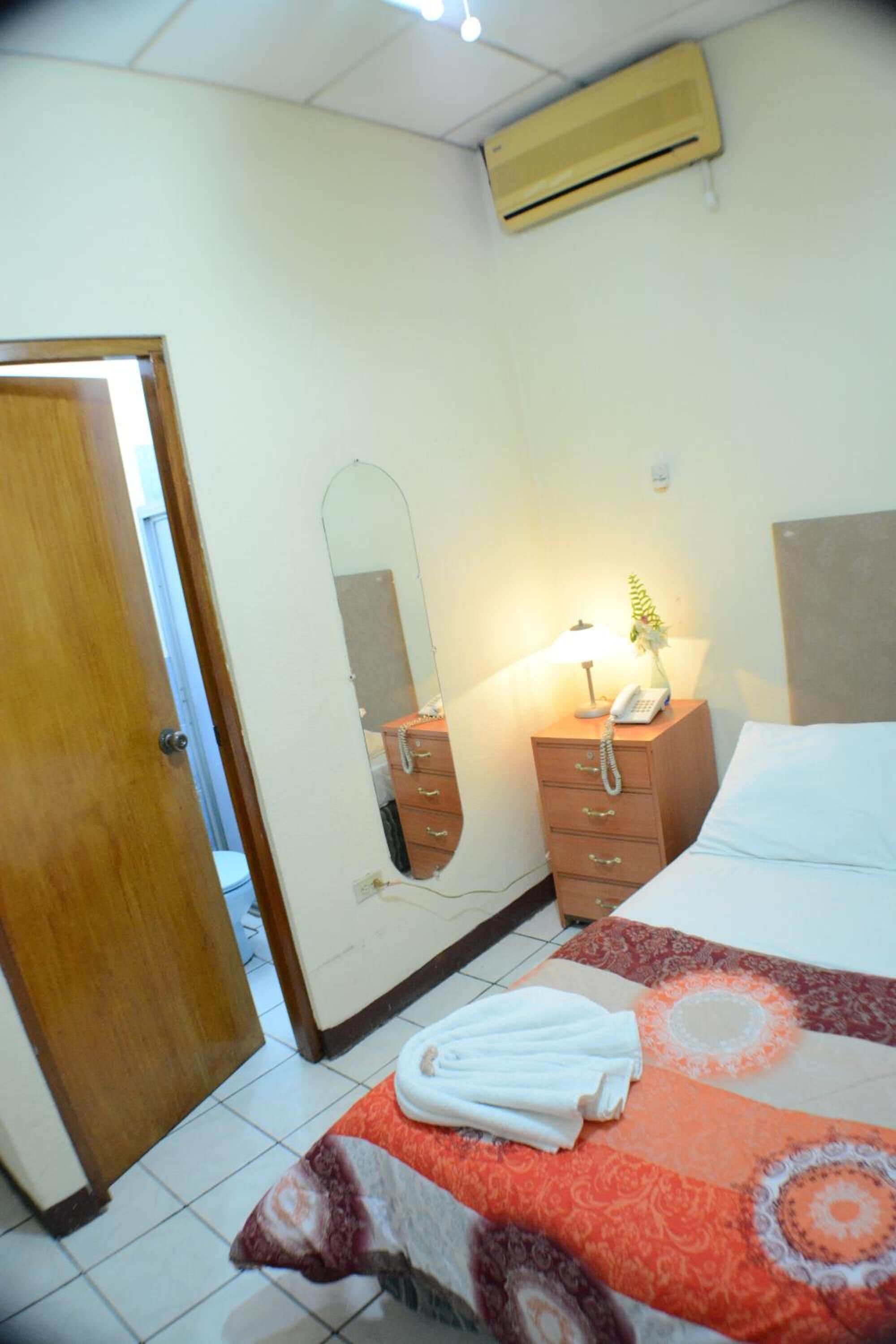 Une chambre d’hôtel comprenant un lit, une table de chevet avec une lampe et un téléphone, une commode, un miroir et un appareil de climatisation.
