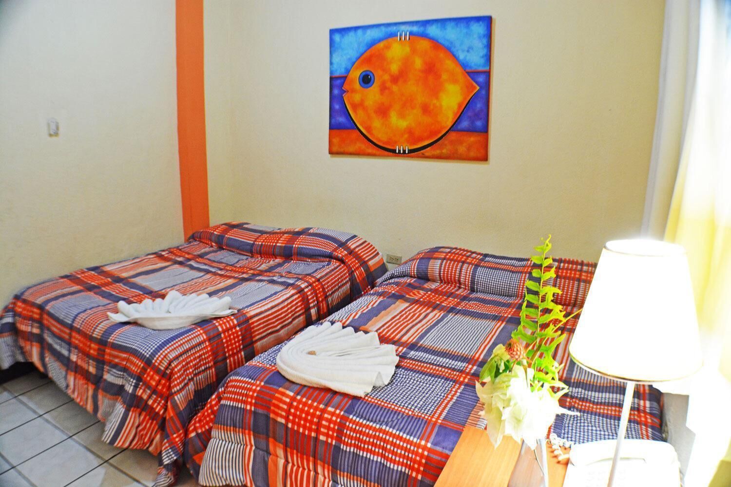 A hotel room with two beds, each with a plaid bedspread and white towels. There is a painting of a fish on the wall and a lamp on the bedside table.