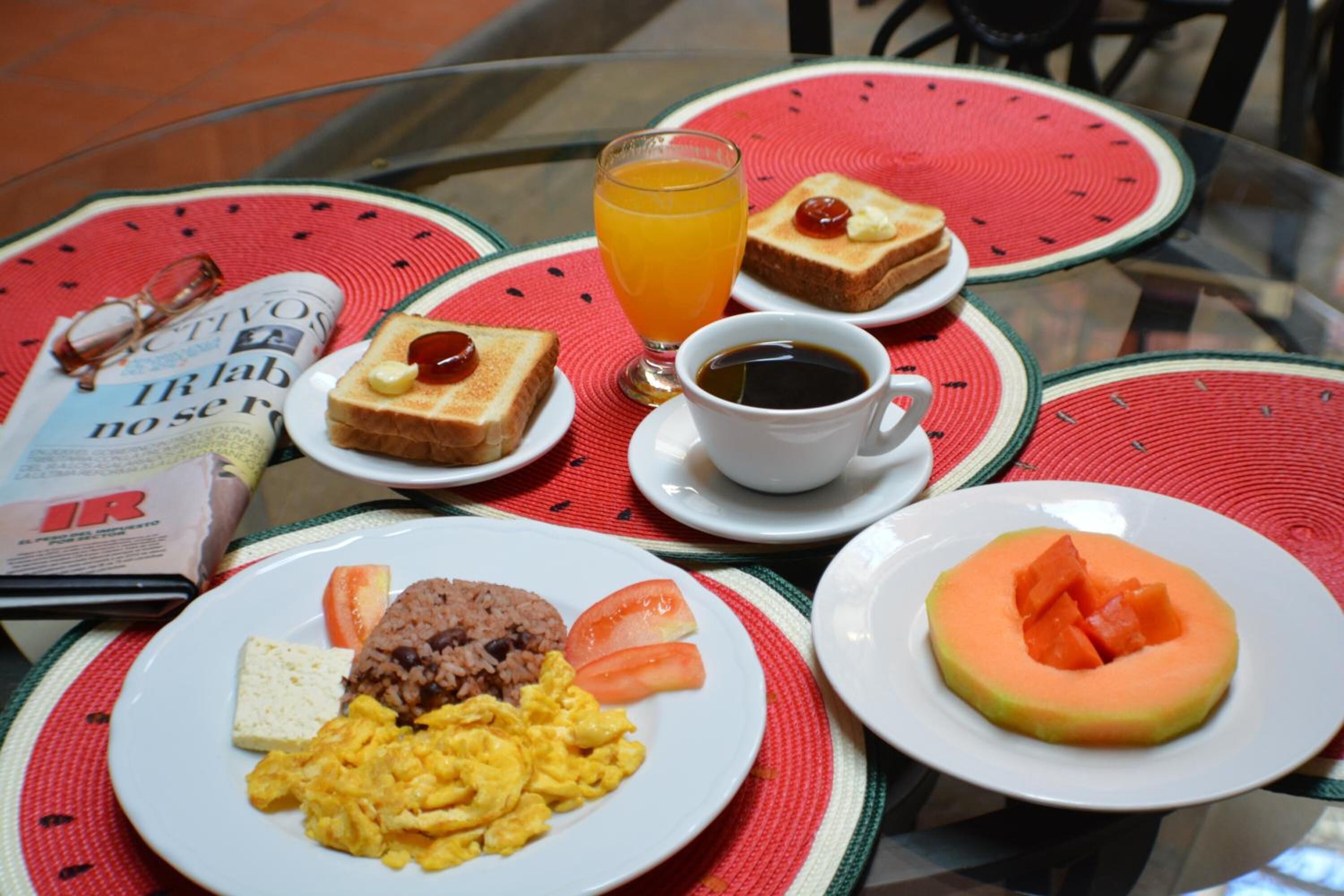 A breakfast setting with toast, scrambled eggs, rice, and a melon slice on a table with a newspaper and a glass of orange juice.