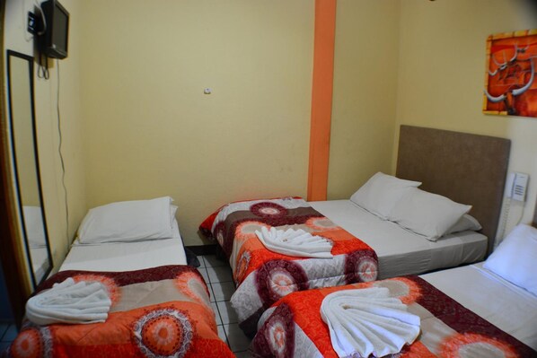 Habitación de hotel con tres camas, cada una con una colcha de cuadros rojos y blancos y almohadas blancas. Hay un televisor montado en la pared y un cuadro sobre la cama de la derecha.