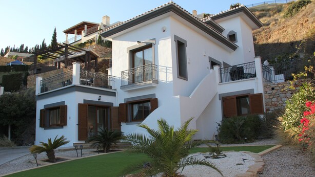 A two-story house with a balcony, a stone retaining wall, and a small garden with palm trees. - Beautiful Detached Spacious Villa, Amazing views, Private Heated Pool, WiFi, A/C (La Manga Club)
