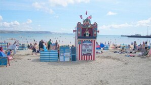 A beach with a Punch & Judy puppet theater, beachgoers, and a pier in the background.