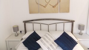 A neatly made bed with a metal headboard, two bedside tables with lamps, and a wall-mounted picture of a beach with heart shapes drawn in the sand.