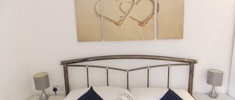 A neatly made bed with a metal headboard, two bedside tables with lamps, and a wall-mounted picture of a beach with heart shapes drawn in the sand.