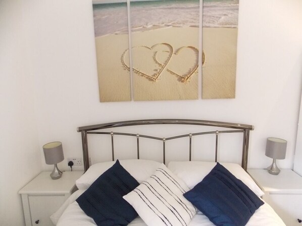A neatly made bed with a metal headboard, two bedside tables with lamps, and a wall-mounted picture of a beach with heart shapes drawn in the sand.