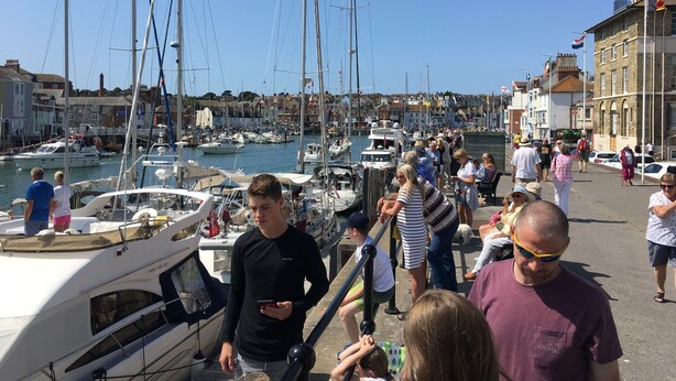 A marina with boats docked, a crowd of people, and buildings in the background.
