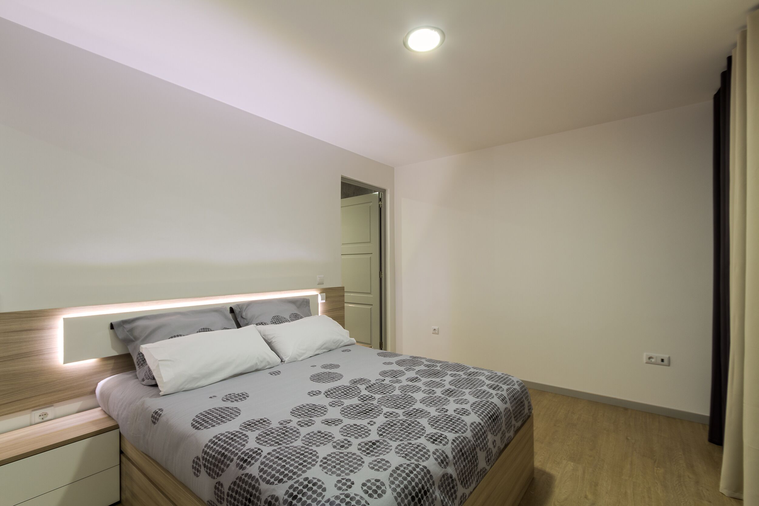 A modern bedroom with a bed, bedside tables, a nightstand, a built-in headboard, and a ceiling light.