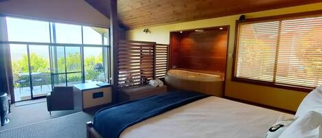 A bedroom with a large bed, a small desk, a window with blinds, a bathtub, and a view of greenery outside.