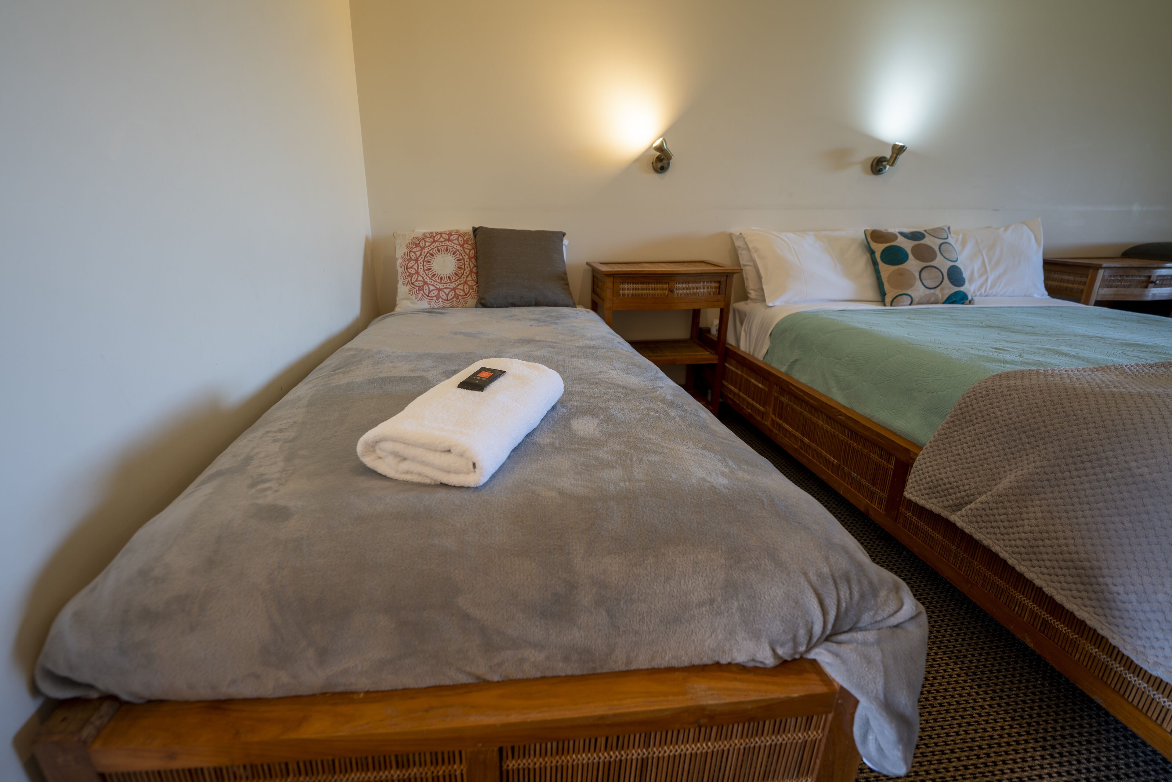 A double bed with a grey blanket and a rolled white towel on the left side. There is a wooden nightstand with a lamp between the beds.