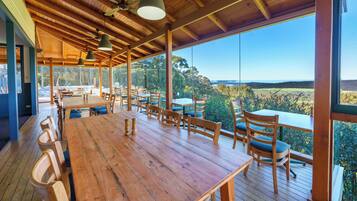 A spacious dining area with wooden tables and chairs, large windows offering a view of the landscape, and ceiling fans.