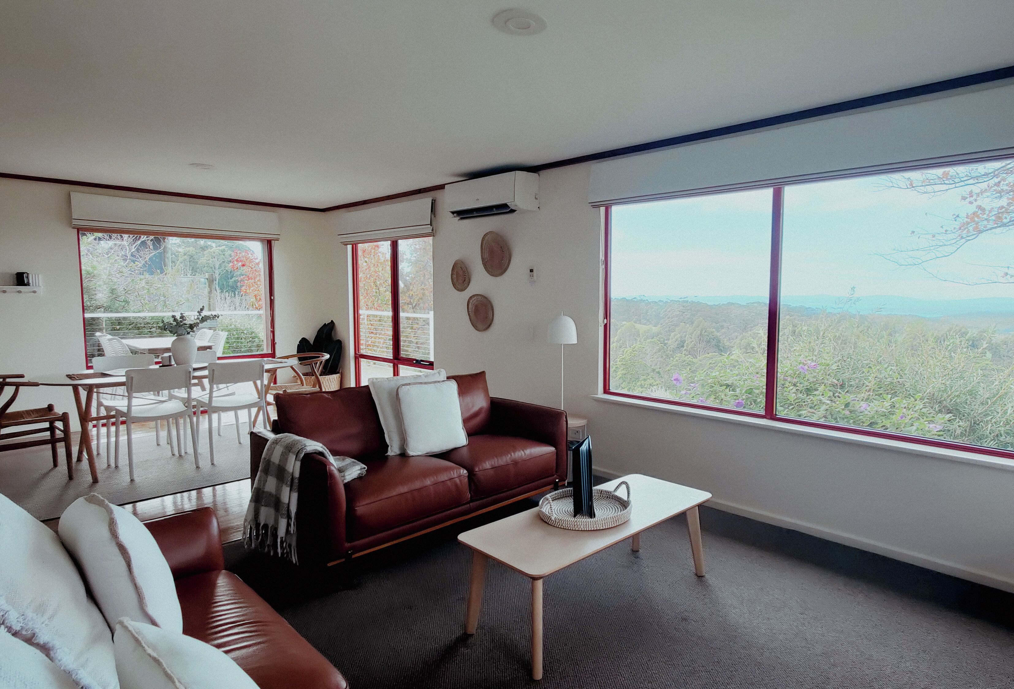 A modern living room with a leather sofa, a coffee table, and a dining area with chairs and a table. Large windows offer a view of the outdoors.