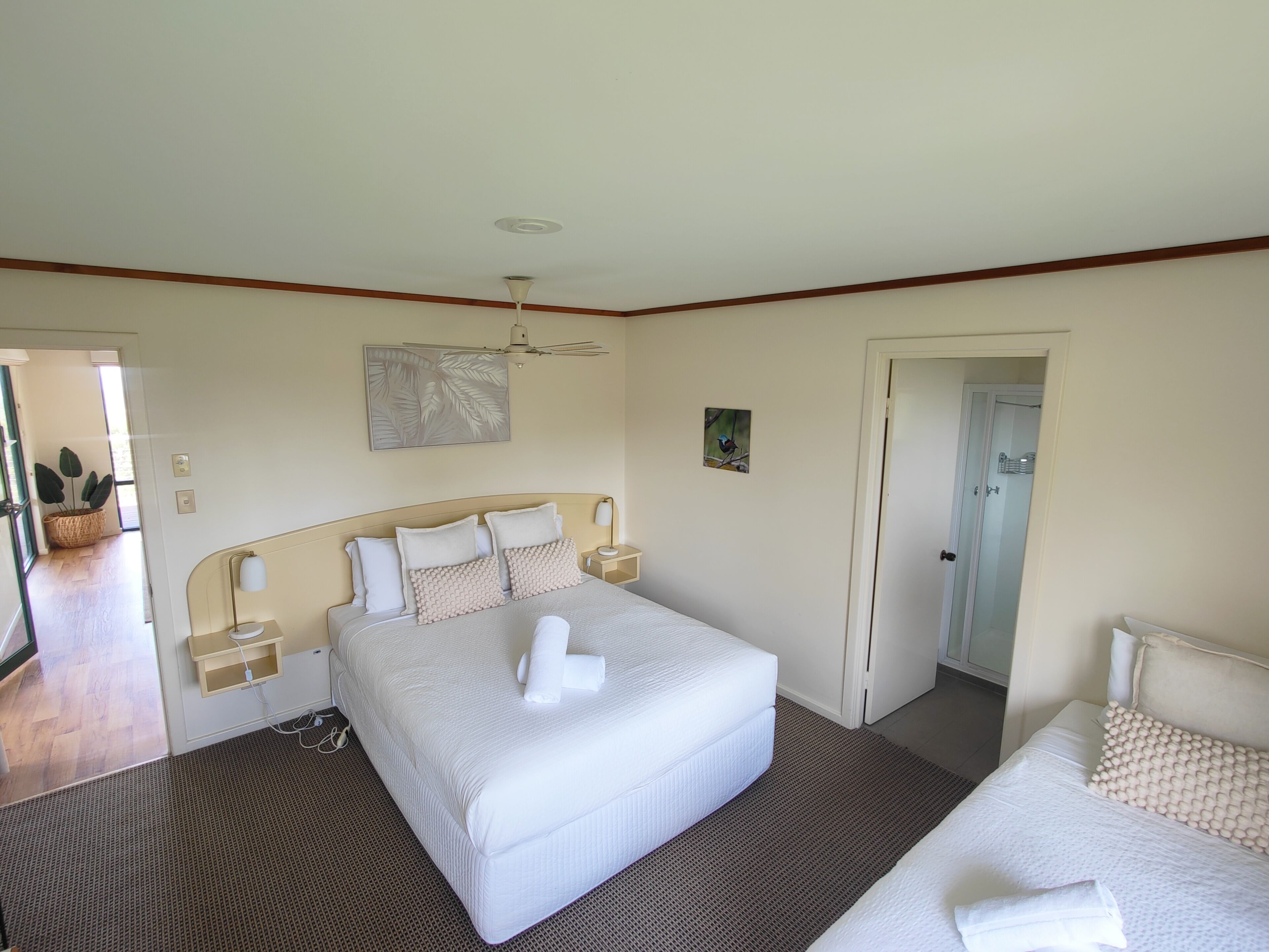 A hotel room with a large bed, bedside tables, a ceiling fan, a painting, and a door leading to another room.