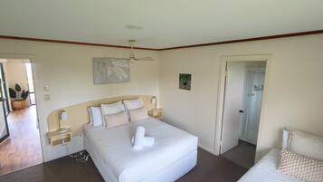 Una habitación de hotel con una cama grande, mesitas de noche, un ventilador de techo, un cuadro y una puerta que da a otra habitación.