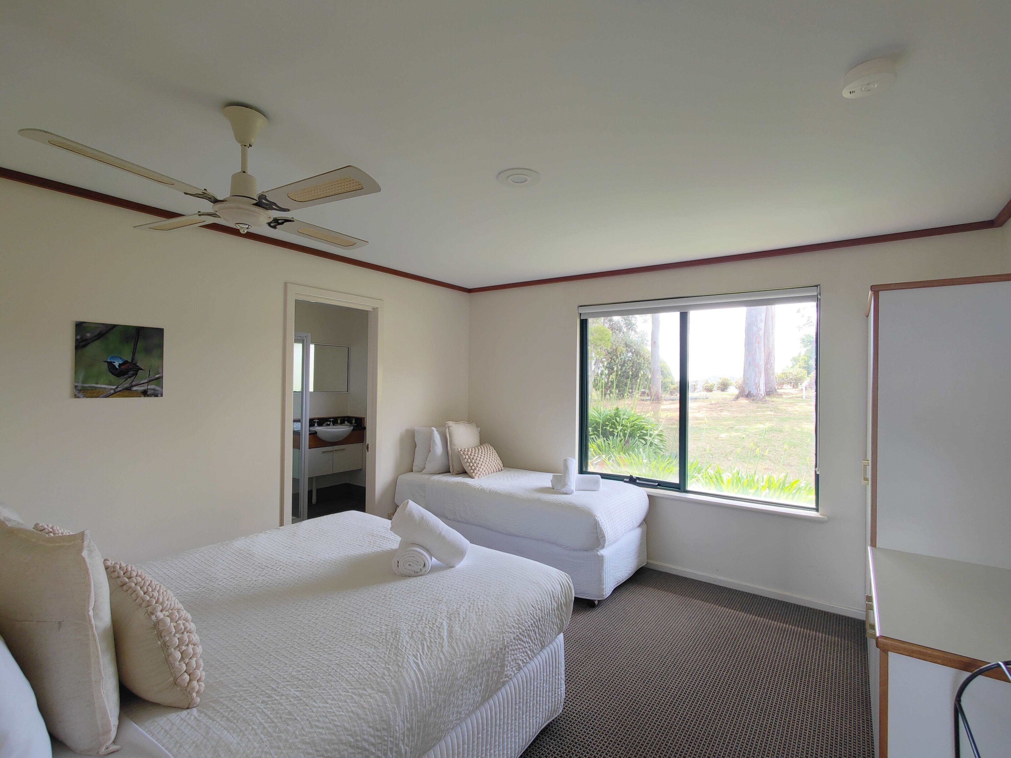 A hotel room with two beds, a ceiling fan, a large window with a view of the outdoors, and a whiteboard on the wall.