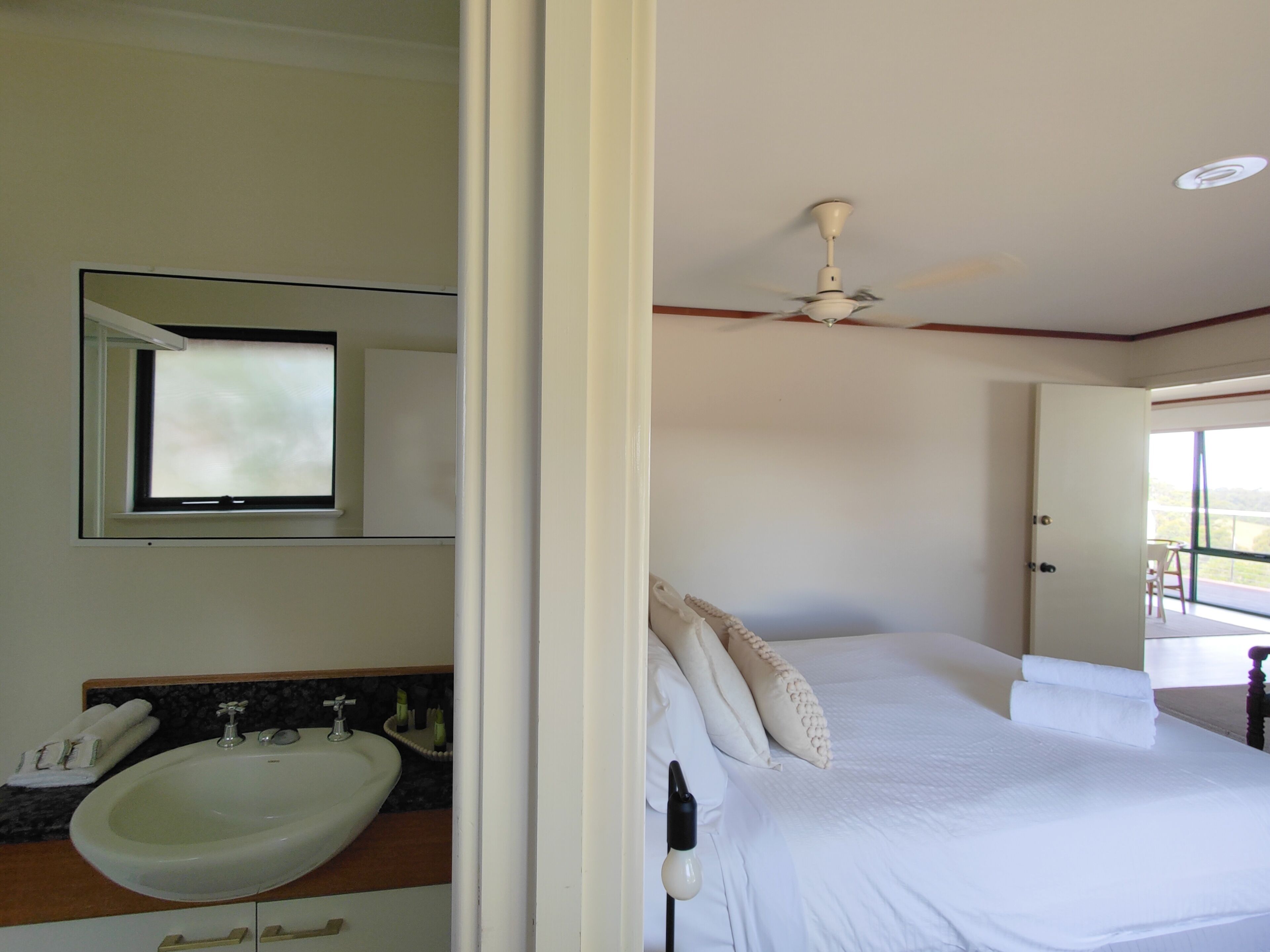 A bedroom with a bed, a ceiling fan, a window, and a bathroom with a sink and mirror.