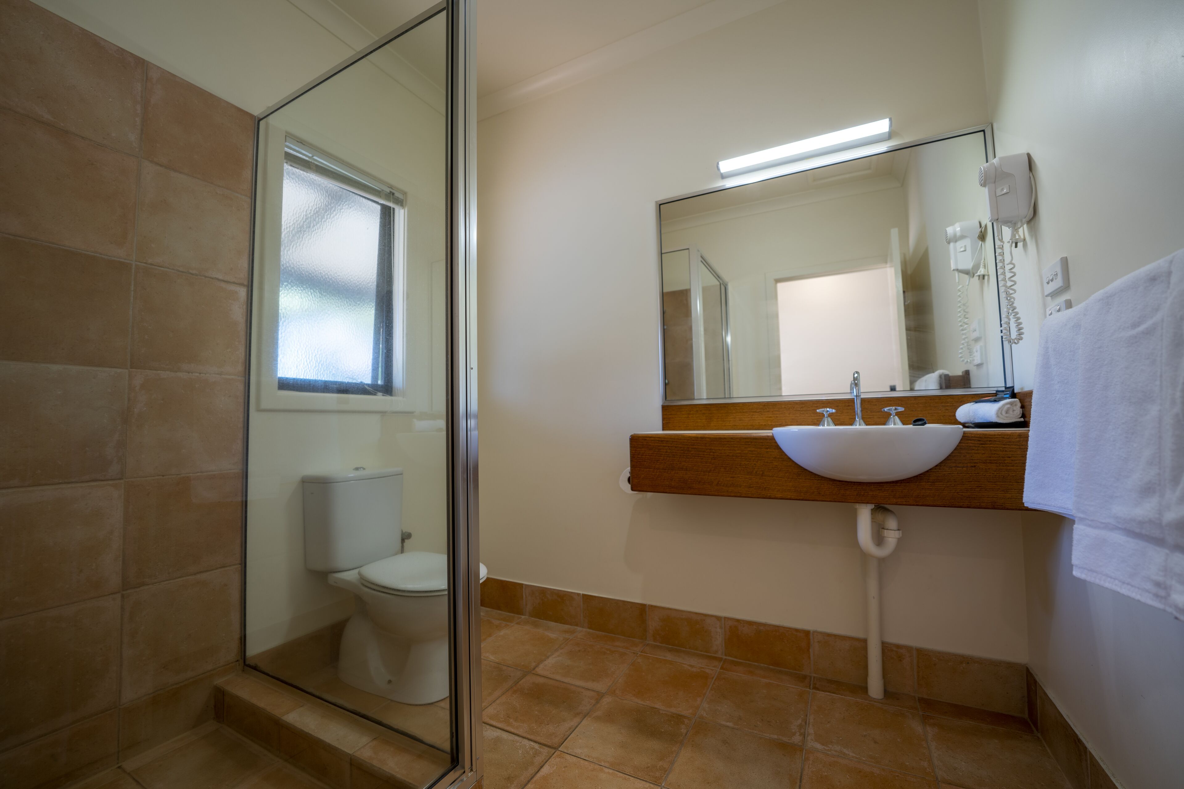 Un baño con cabina de ducha de vidrio, inodoro blanco, tocador de madera con lavamanos blanco, espejo y toalla colgada en la pared.