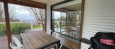Une terrasse couverte avec une table en bois et des chaises blanches, un barbecue et une vue sur l’extérieur.