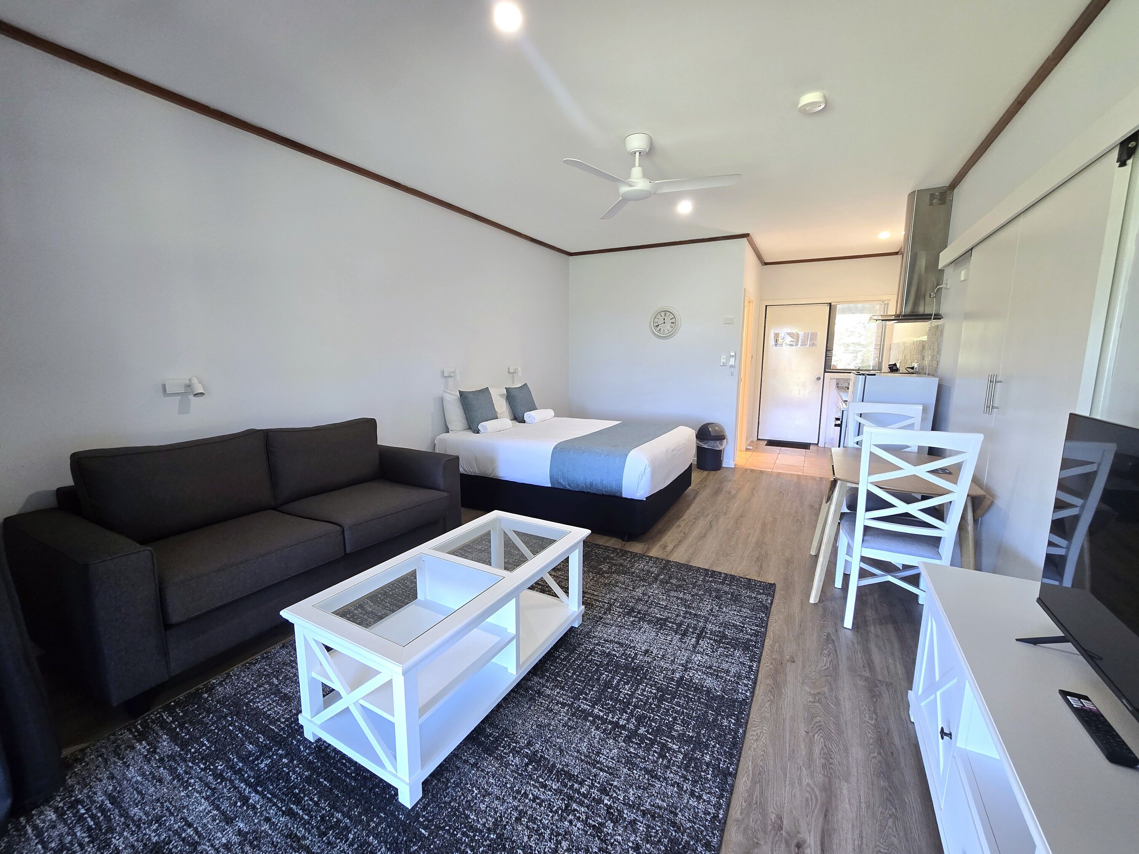 A modern hotel room with a dark grey sofa, a bed with white linens, a white coffee table, and a small kitchen area with a microwave and a kettle.