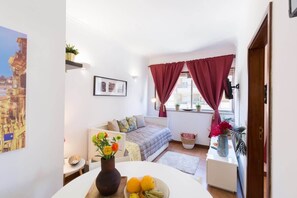 Una sala de estar acogedora con un sofá, una mesa de comedor con fruta, un florero con flores y una ventana con cortinas.