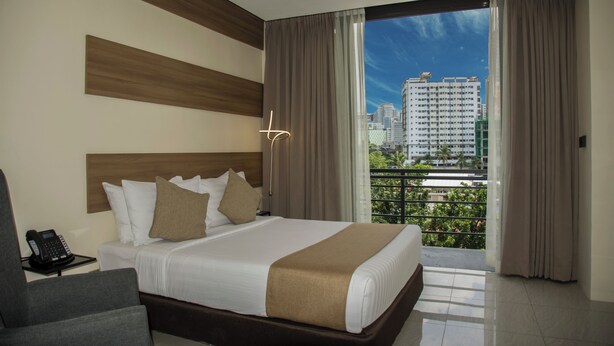 A hotel room with a large bed, a bedside phone, a chair, and a view of the city through a balcony.