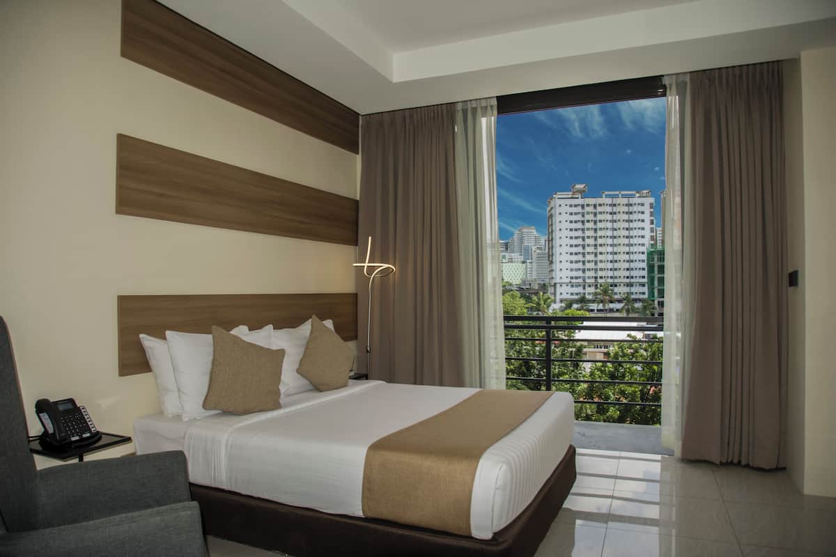 A hotel room with a large bed, a bedside phone, a chair, and a view of the city through a balcony.