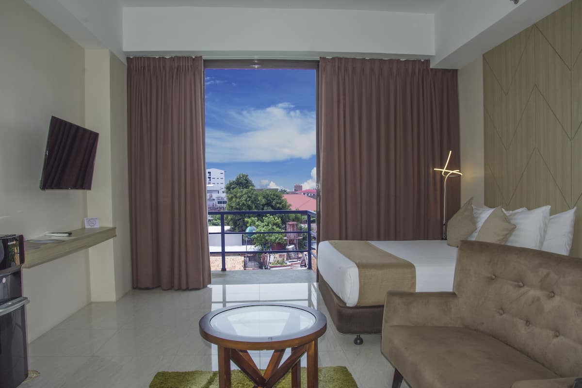 A hotel room with a bed, a sofa, a small table, a TV, and a balcony with curtains.